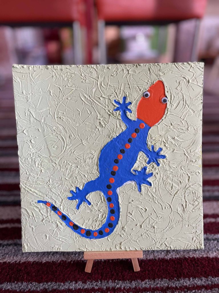 Schilderij van een blauwe gekko met oranje kop op een crèmekleurig doek met reliëf